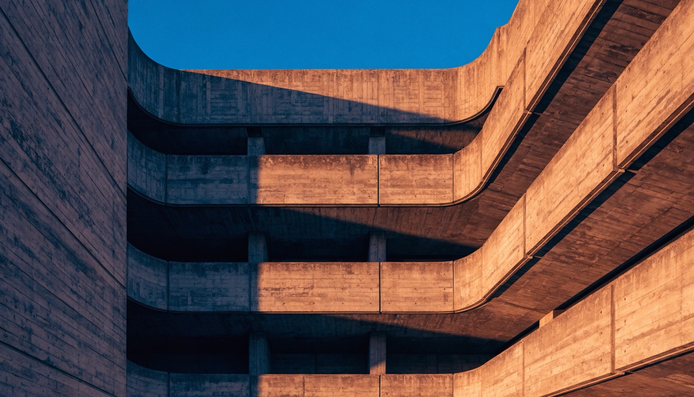 Photograph of a brutalist concrete building at golden hour, board formed balconies curving around a corner in stacked tiers, warm amber sunlight raking across the facade with deep shadow beneath each slab, against a clear cobalt sky