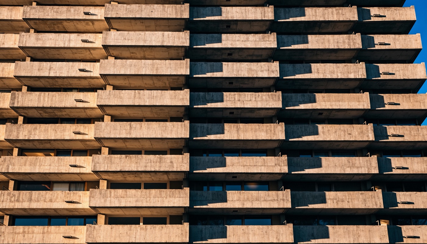 Photograph of a board-marked concrete apartment facade, rows of deep balconies forming a grid, hard low golden sunlight throwing long shadows and a strip of deep blue sky at the top right corner
