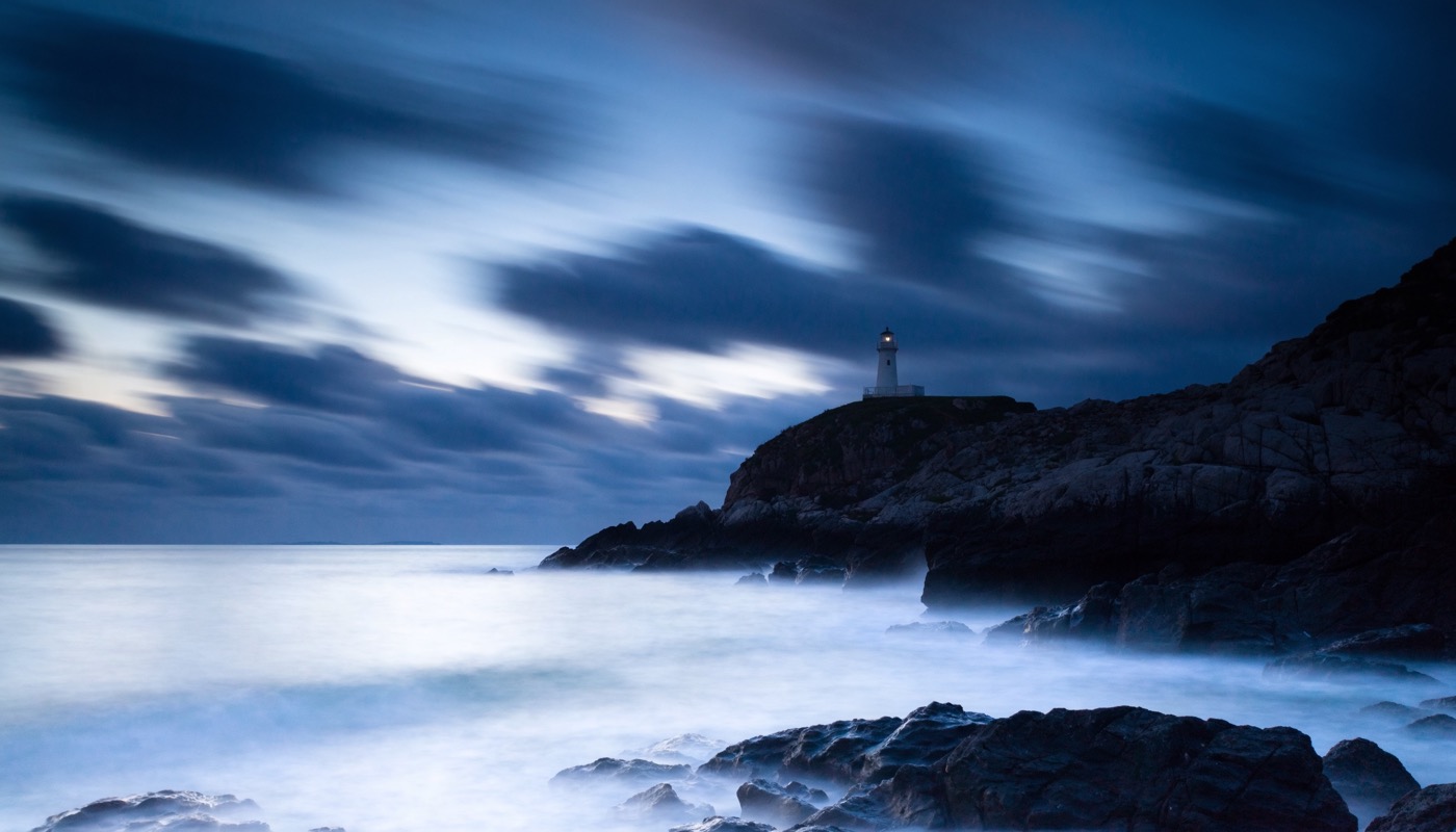 Photographic image of a lighthouse at blue hour, one lamp lit against the weather