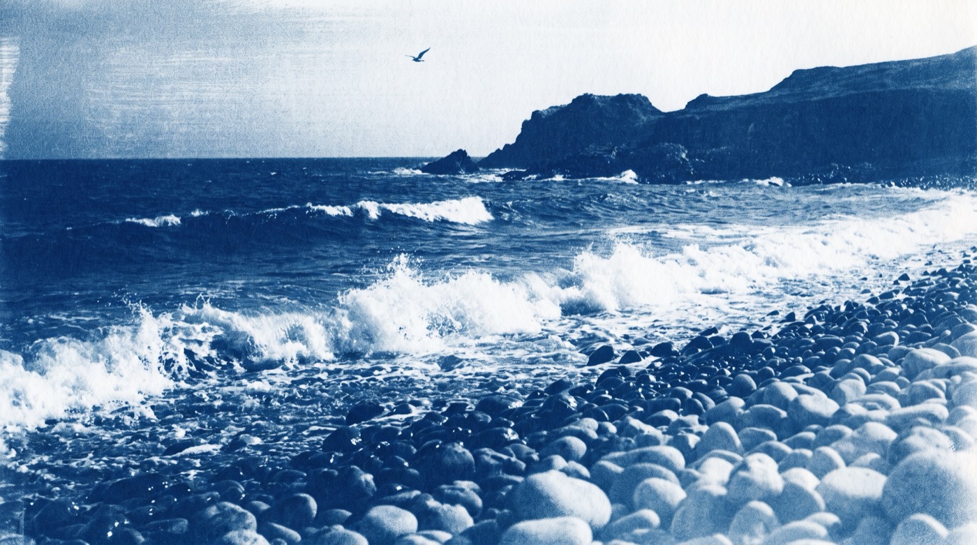 Cyanotype print in deep Prussian blue of white waves breaking on a steep shore of rounded pebbles, a dark headland on the horizon and a single seabird in a pale sky