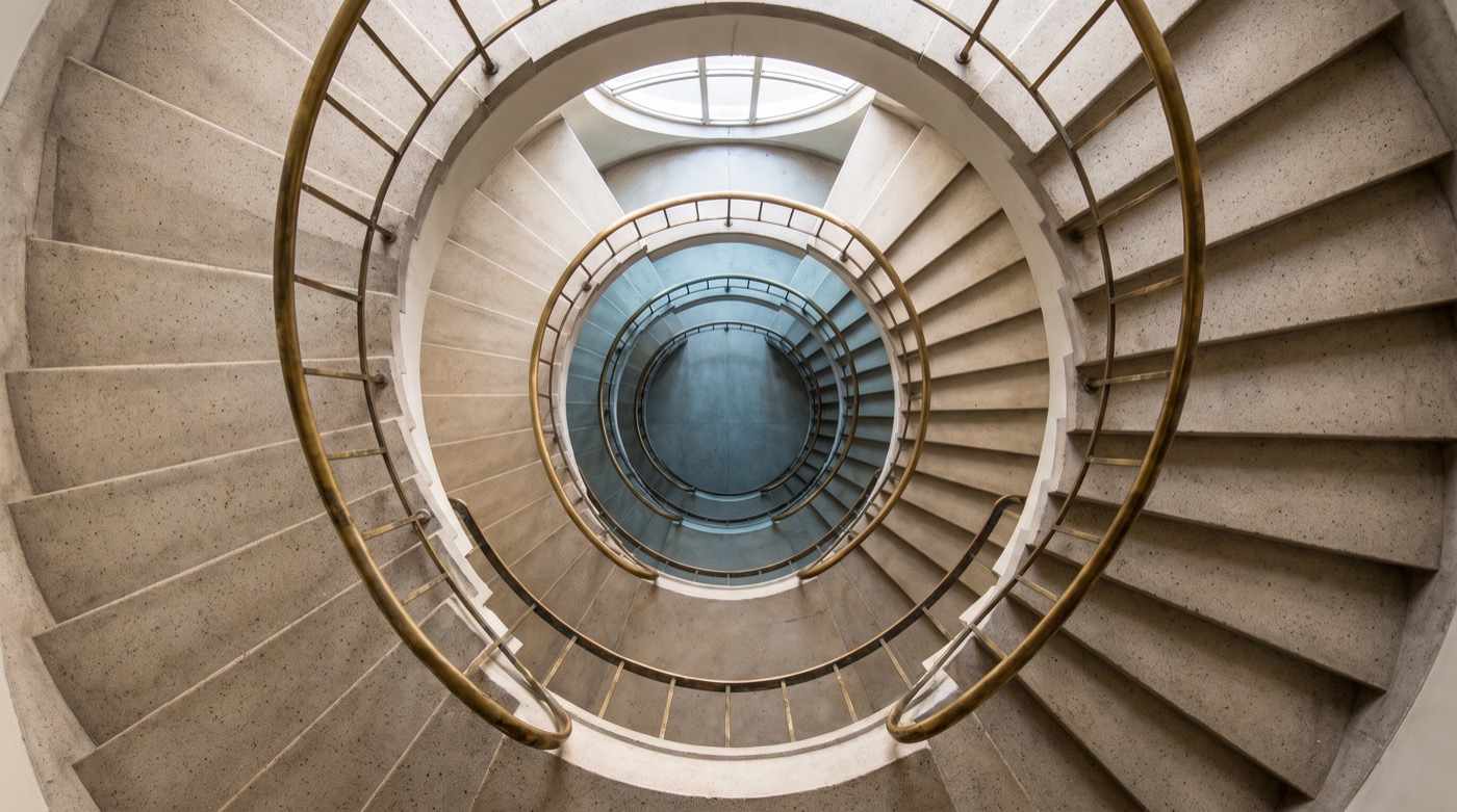 Photograph looking straight down the well of a pale concrete spiral staircase, a slim brass handrail tracing each turn toward a cool blue core, soft daylight falling from a round skylight