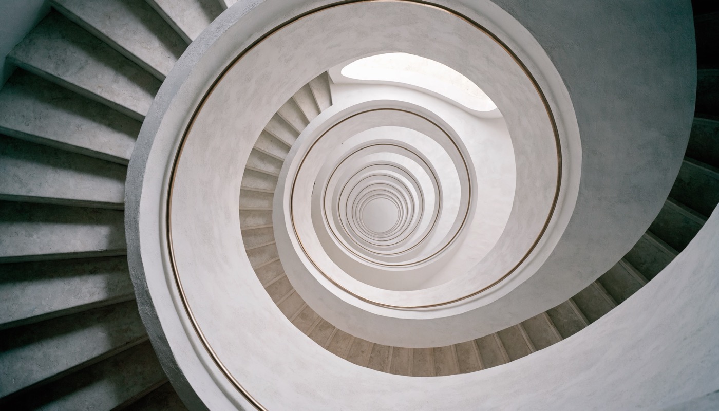 Photograph looking straight down a spiral staircase, pale stone treads and smooth white plaster curving into a deep descending helix with a slender brass handrail, lit softly from a skylight above